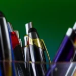 A stock image of a selection of ballpoint pens in a glass or plastic see through holder with a green and blue blended background.
