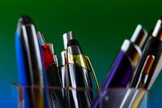 A stock image of a selection of ballpoint pens in a glass or plastic see through holder with a green and blue blended background.
